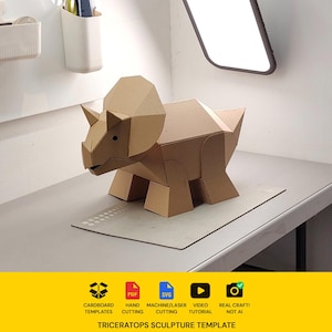 May include: A geometric triceratops sculpture constructed from brown cardboard, featuring a large frill, three horns, and a segmented body. Below the sculpture, a yellow banner displays icons for cardboard templates, PDF, SVG, video tutorial, and real craft.