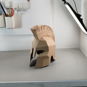 May include: A tan cardboard Spartan-style helmet featuring a prominent crest along the top, a T-shaped face opening, and angular geometric construction. The helmet is crafted from layered brown corrugated paper material with visible seams.