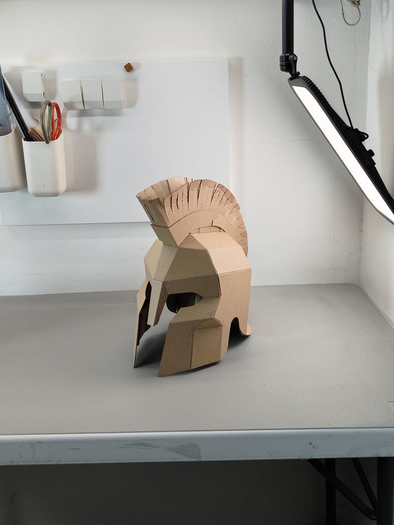 May include: A tan cardboard Spartan-style helmet featuring a prominent crest along the top, a T-shaped face opening, and angular geometric construction. The helmet is crafted from layered brown corrugated paper material with visible seams.