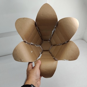 May include: A bowl-shaped structure constructed from brown corrugated cardboard, featuring six large, petal-like segments that curve outward from a central hexagonal base. The edges of the cardboard pieces are joined with visible white adhesive.
