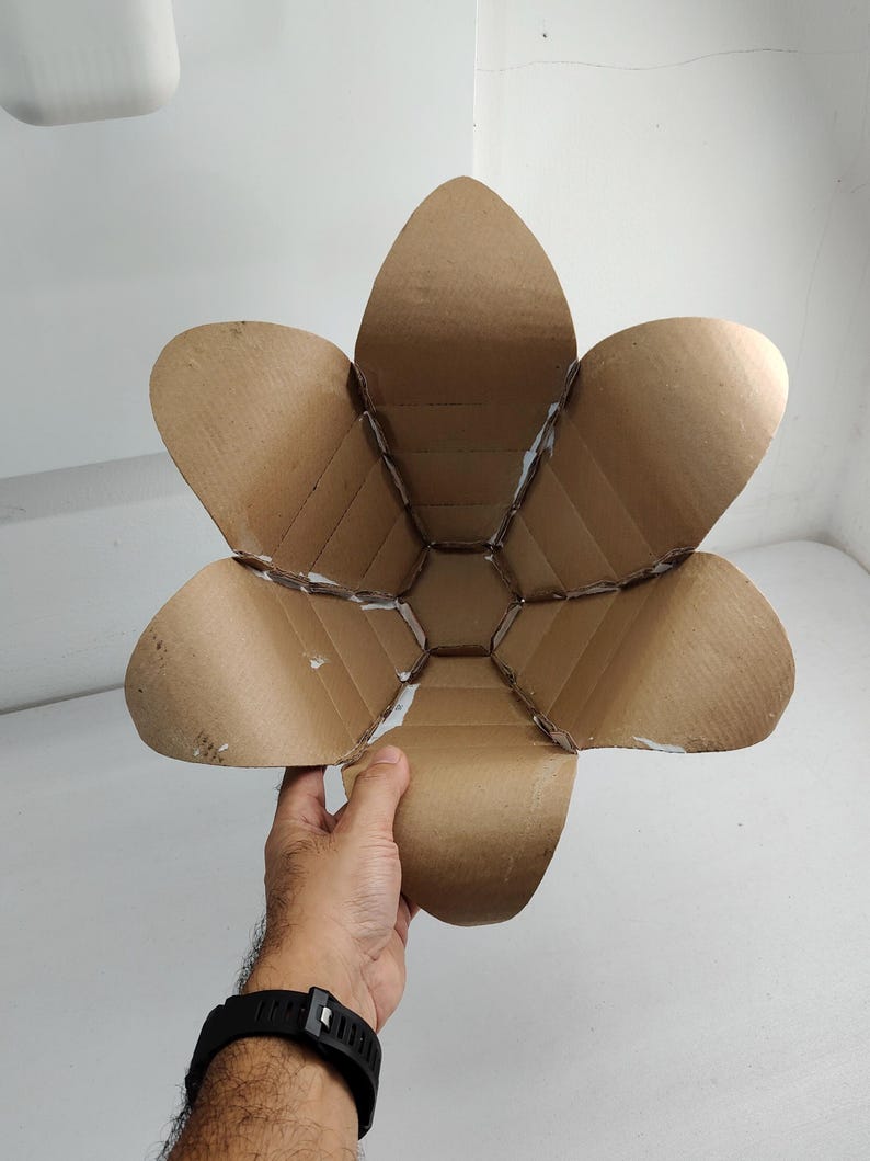 May include: A bowl-shaped structure constructed from brown corrugated cardboard, featuring six large, petal-like segments that curve outward from a central hexagonal base. The edges of the cardboard pieces are joined with visible white adhesive.