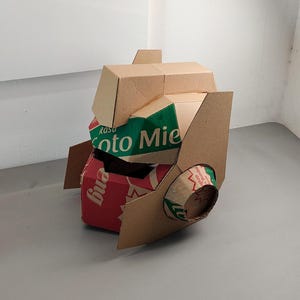 May include: A sculptural helmet constructed from repurposed cardboard packaging. The structure features tan, red, and green panels with visible text including 'Rasa' and 'Soto Mie'. It has a geometric, angular design with a cylindrical side projection.