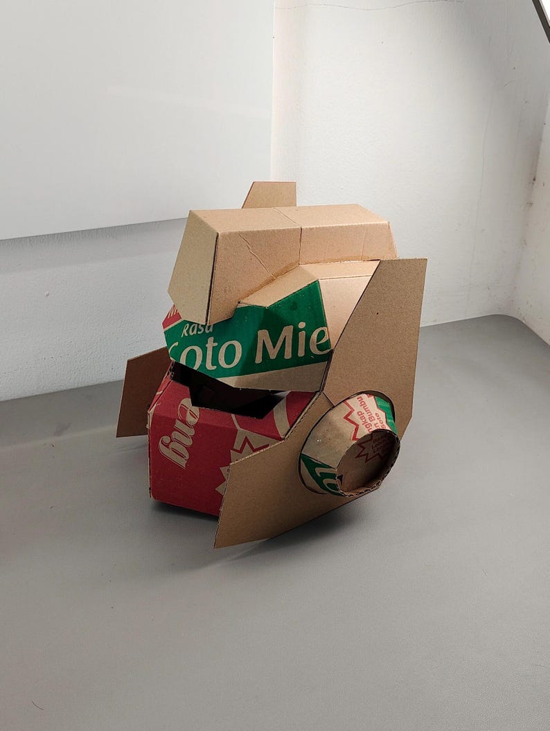 May include: A sculptural helmet constructed from repurposed cardboard packaging. The structure features tan, red, and green panels with visible text including 'Rasa' and 'Soto Mie'. It has a geometric, angular design with a cylindrical side projection.