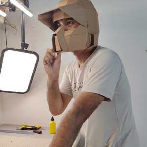 Op de afbeelding: Een geometrische, hoekige helm gemaakt van bruin karton. De helm is voorzien van een opvallend vizier, een gestructureerde gezichtsbeschermer met een centrale opening en een veelzijdig ontwerp dat het hoofd en het gezicht bedekt.