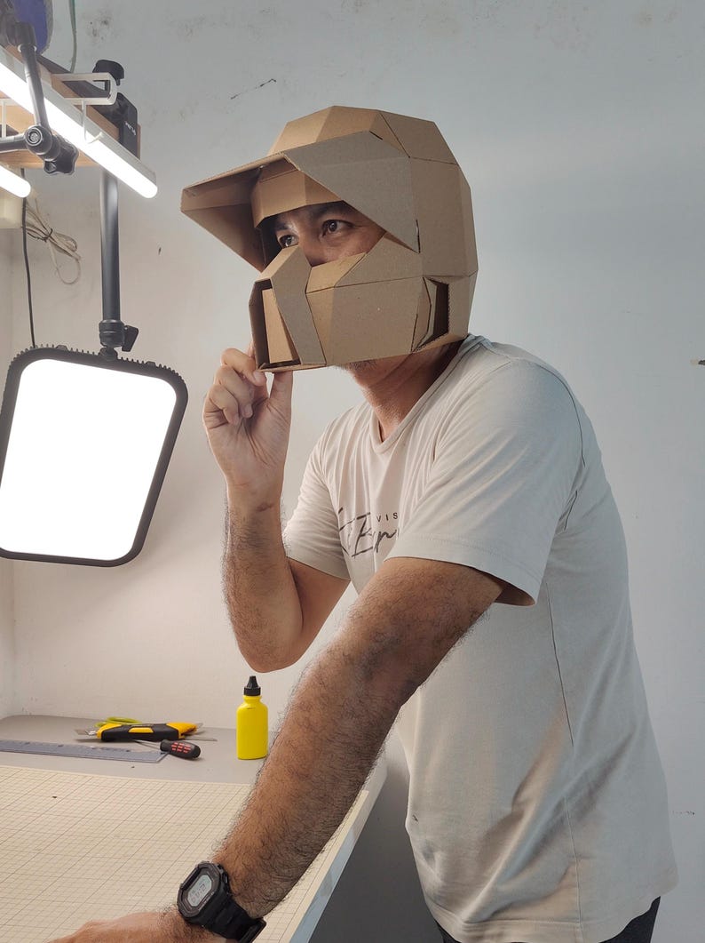 Op de afbeelding: Een geometrische, hoekige helm gemaakt van bruin karton. De helm is voorzien van een opvallend vizier, een gestructureerde gezichtsbeschermer met een centrale opening en een veelzijdig ontwerp dat het hoofd en het gezicht bedekt.