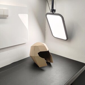 May include: A geometric helmet constructed from tan cardboard, featuring a faceted, angular design with a wide, open visor area. The helmet is composed of multiple flat, interconnected panels that form a structured, three-dimensional shape.