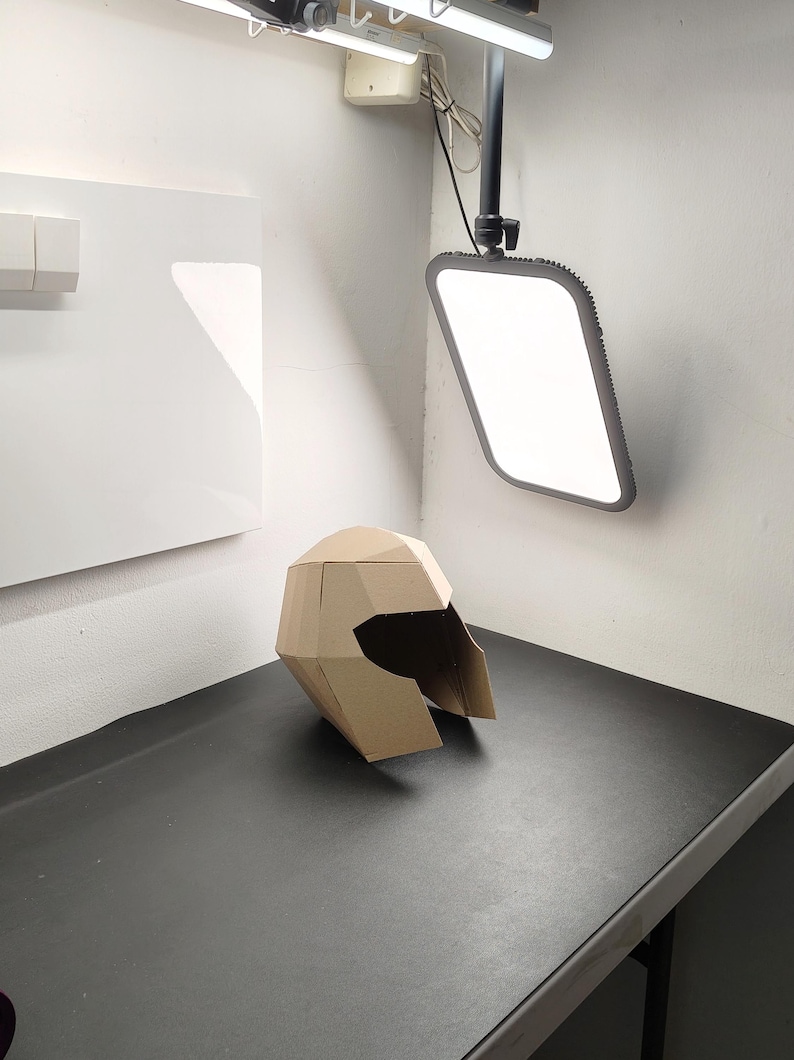 May include: A geometric helmet constructed from tan cardboard, featuring a faceted, angular design with a wide, open visor area. The helmet is composed of multiple flat, interconnected panels that form a structured, three-dimensional shape.