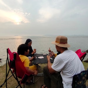 Op de afbeelding: Vier mensen zitten aan een tafel en spelen kaarten. De tafel is van hout en heeft een zwart blad. De mensen zitten op stoelen. Er is een strand op de achtergrond.