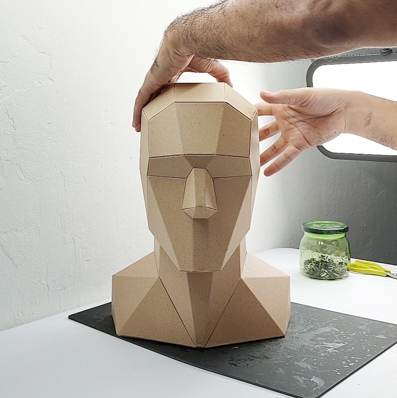 May include: A brown cardboard model of a human head and neck, with geometric shapes forming the features. The model is being held by a hand.