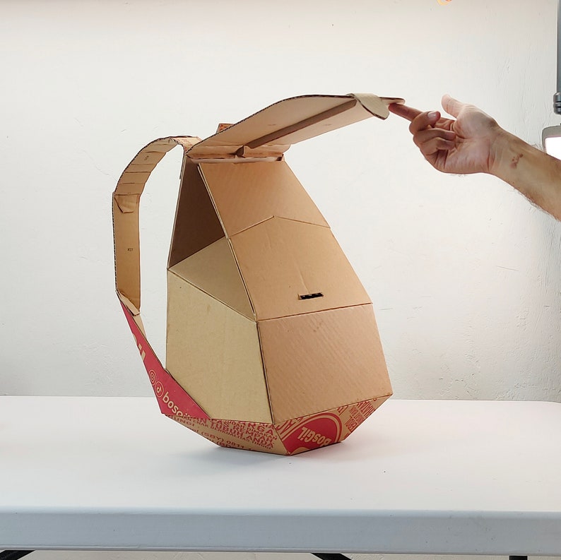 May include: A cardboard backpack with a flap that opens up. The backpack is made from recycled cardboard and has a red and white pattern on the bottom.