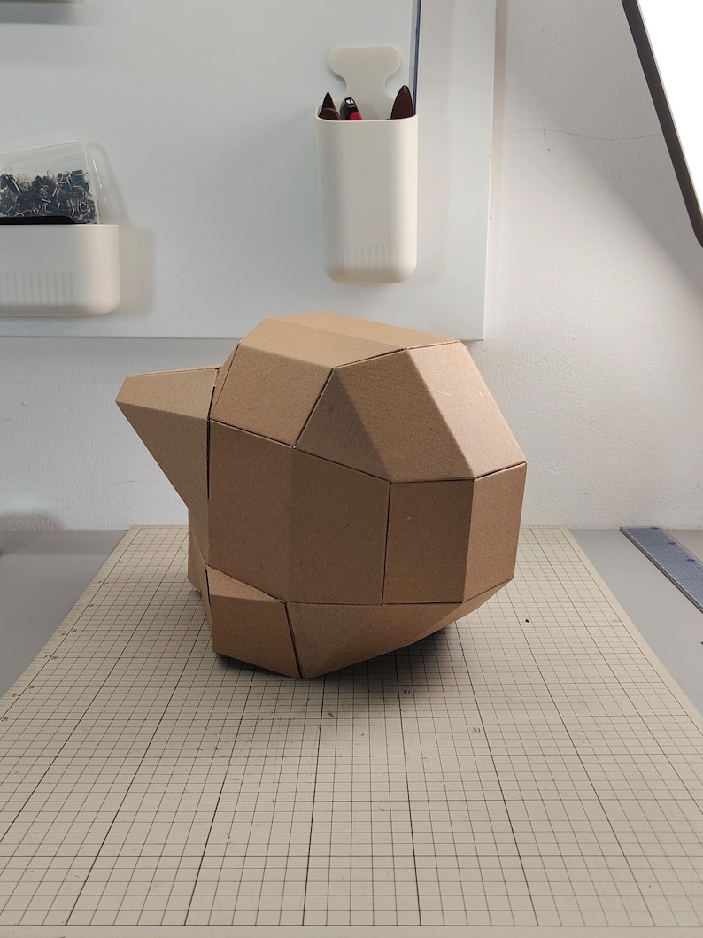 Op de afbeelding: Een geometrisch, driedimensionaal beeldhouwwerk opgebouwd uit lichtbruine kartonnen panelen. Het object heeft een gefacetteerd, hoekig ontwerp met meerdere platte oppervlakken die samen een complexe, afgeronde en asymmetrische vorm vormen.