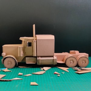 Pode incluir: Um modelo de cami&atilde;o de plataforma com uma cabine e um reboque castanhos, feito de cart&atilde;o canelado. O modelo tem um design detalhado. O cami&atilde;o est&aacute; sobre uma superf&iacute;cie verde.
