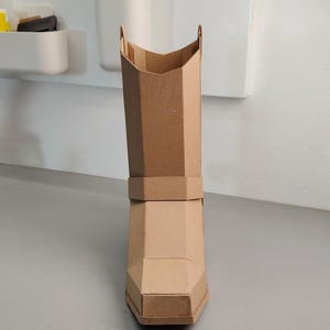 Puede incluir: Una bota alta y estructurada hecha de cart&oacute;n marr&oacute;n claro. La bota presenta un dise&ntilde;o geom&eacute;trico y facetado con punta cuadrada, una banda en la parte media de la ca&ntilde;a y una abertura en forma de V en la parte superior de la ca&ntilde;a.