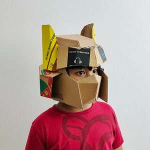 Peut inclure: Un enfant portant un casque de robot en carton fabriqué à partir d'emballages alimentaires recyclés. Le casque est brun et jaune avec un motif bleu et blanc. Le casque a une visière et une paire d'écouteurs sont visibles à l'intérieur.