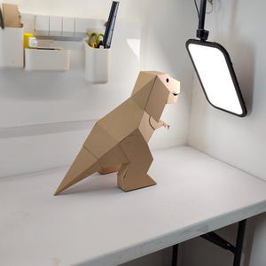 May include: A geometric dinosaur figure constructed from tan cardboard. The sculpture features a faceted, angular design with a long tail, small arms, and a distinct head shape, standing in a profile view with a visible eye and mouth detail.