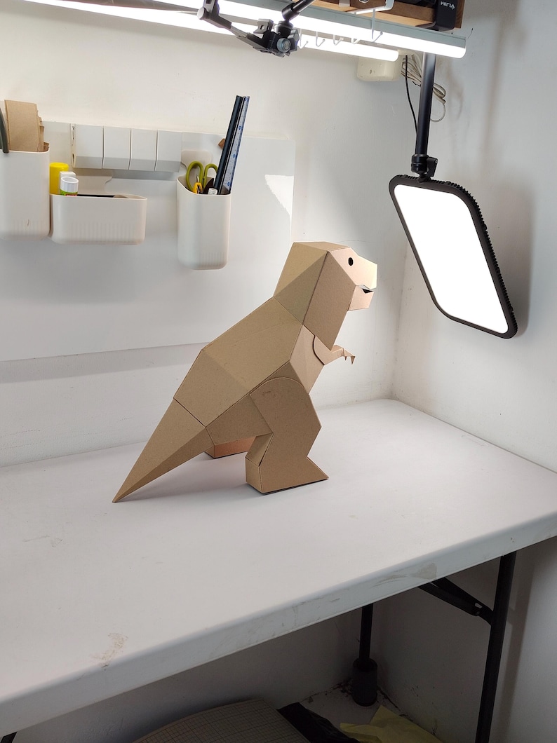 May include: A geometric dinosaur figure constructed from tan cardboard. The sculpture features a faceted, angular design with a long tail, small arms, and a distinct head shape, standing in a profile view with a visible eye and mouth detail.