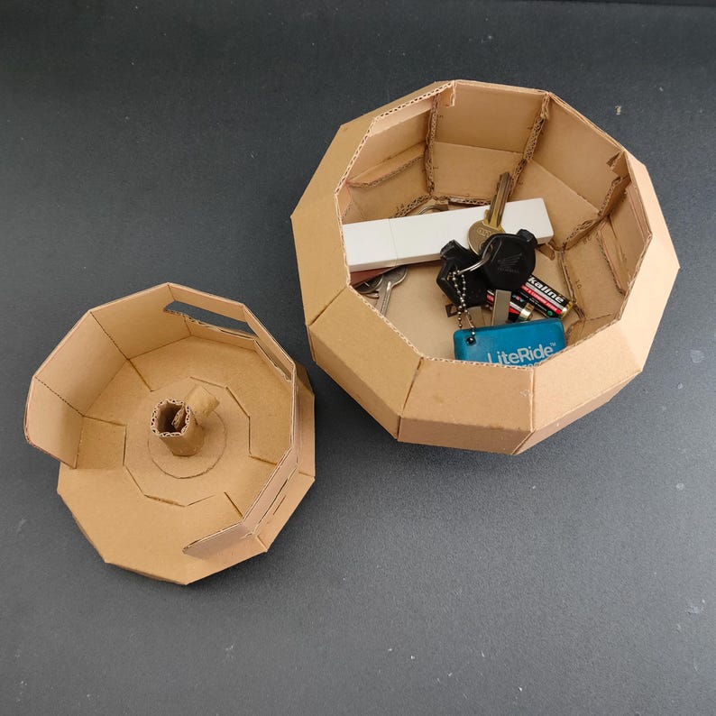 May include: An octagonal cardboard storage container with a matching lid. The open container holds a white USB drive, a set of metal keys on a ring, two AA batteries, and a small blue rectangular object with the text LiteRide printed on it.