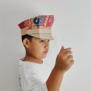 May include: A handmade cardboard hat, resembling a military cap, is worn by a young person. The hat is constructed from repurposed cardboard packaging, featuring red, brown, and white printed text. The brim and crown are distinct, showcasing a creative DIY project.