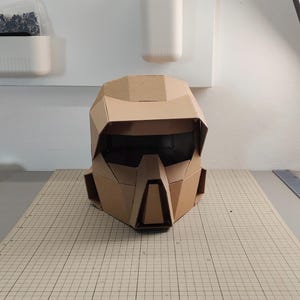 Op de afbeelding: Een geometrische helm gemaakt van bruin golfkarton. De helm heeft een brede vizieropening, een prominente centrale neusbeschermer en hoekige zijpanelen. Het ontwerp is samengesteld uit meerdere platte, gevouwen kartonnen segmenten.