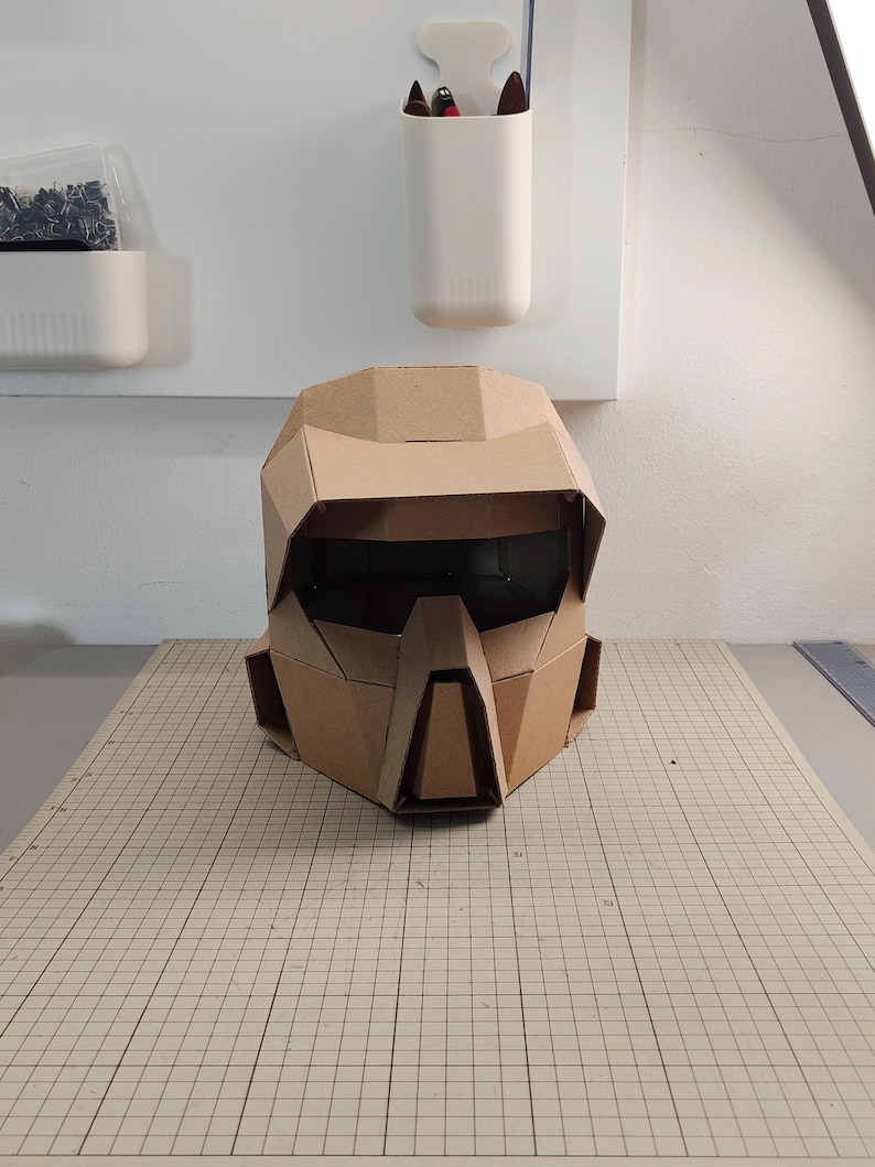 Op de afbeelding: Een geometrische helm gemaakt van bruin golfkarton. De helm heeft een brede vizieropening, een prominente centrale neusbeschermer en hoekige zijpanelen. Het ontwerp is samengesteld uit meerdere platte, gevouwen kartonnen segmenten.
