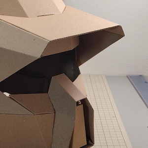 Op de afbeelding: Een geometrische helm gemaakt van bruin golfkarton, voorzien van een breed vizier, hoekige gezichtsplaten en een centrale kinbeschermer. De structuur bestaat uit meerdere gevouwen panelen die samen een stijve hoofdbedekking vormen.
