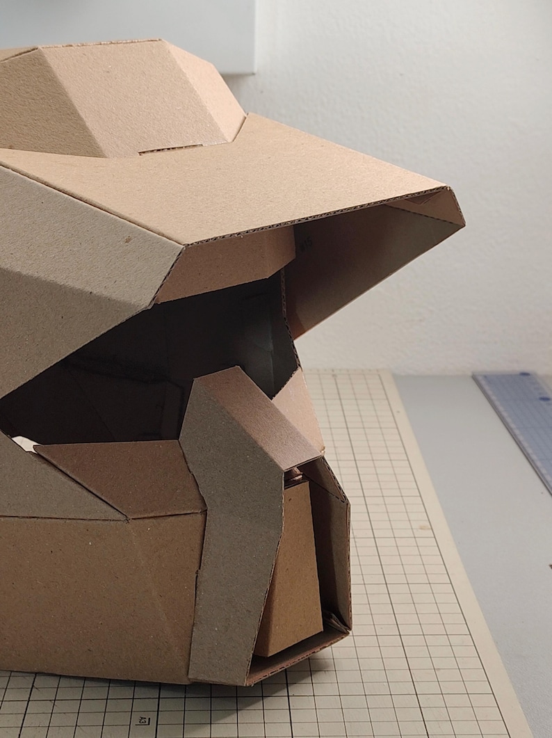 Op de afbeelding: Een geometrische helm gemaakt van bruin golfkarton, voorzien van een breed vizier, hoekige gezichtsplaten en een centrale kinbeschermer. De structuur bestaat uit meerdere gevouwen panelen die samen een stijve hoofdbedekking vormen.