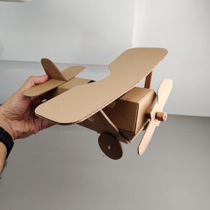May include: A handcrafted biplane model constructed from brown corrugated cardboard. The aeroplane features two stacked wings, a central fuselage, a front-mounted propeller, and circular wheels attached to the sides of the main body structure.