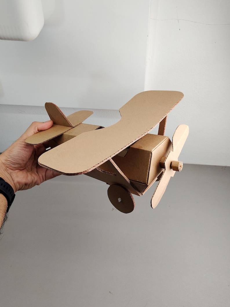 May include: A handcrafted biplane model constructed from brown corrugated cardboard. The aeroplane features two stacked wings, a central fuselage, a front-mounted propeller, and circular wheels attached to the sides of the main body structure.