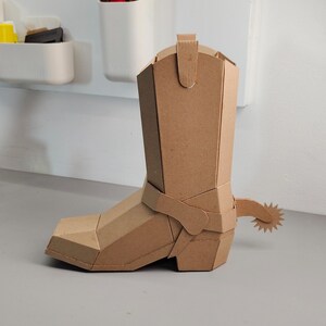 Puede incluir: Una escultura tridimensional de bota vaquera construida con cart&oacute;n marr&oacute;n. La bota presenta un dise&ntilde;o geom&eacute;trico facetado con punta cuadrada, una ca&ntilde;a alta con tirador y una espuela decorativa unida al tal&oacute;n con una rodaja en forma de estrella.