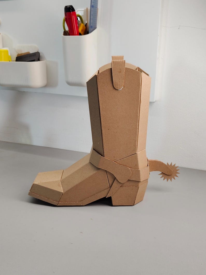 Puede incluir: Una escultura tridimensional de bota vaquera construida con cart&oacute;n marr&oacute;n. La bota presenta un dise&ntilde;o geom&eacute;trico facetado con punta cuadrada, una ca&ntilde;a alta con tirador y una espuela decorativa unida al tal&oacute;n con una rodaja en forma de estrella.