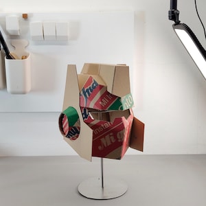 May include: A helmet sculpture constructed from recycled cardboard packaging featuring red, green, and white branding with the visible text Indomie, Mi Instan, and Soto. The helmet is mounted on a silver metal stand with a circular base.
