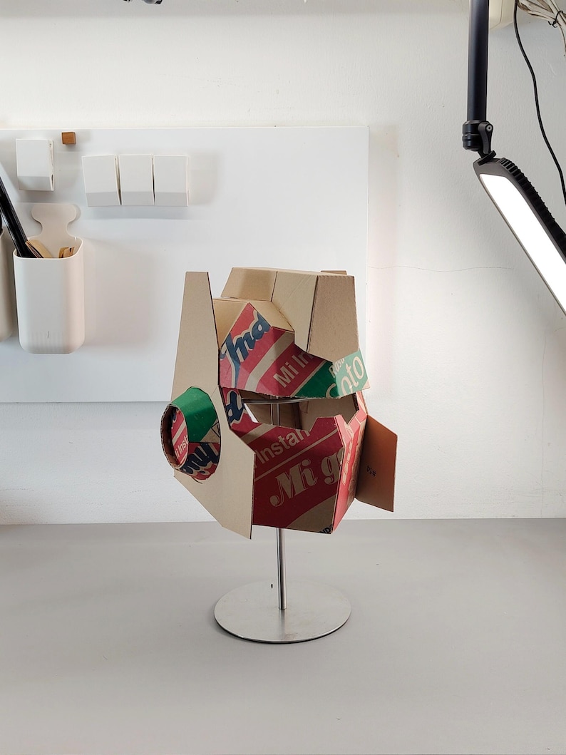 May include: A helmet sculpture constructed from recycled cardboard packaging featuring red, green, and white branding with the visible text Indomie, Mi Instan, and Soto. The helmet is mounted on a silver metal stand with a circular base.