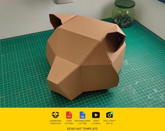 May include: A geometric bear head sculpture constructed from brown cardboard with angular facets and two pointed ears. Below the object, a yellow banner displays the text: CARDBOARD TEMPLATES, PDF, SVG, HAND CUTTING, MACHINE/LASER CUTTING, VIDEO TUTORIAL, REAL CRAFT! NOT AI, BEAR HAT TEMPLATE.