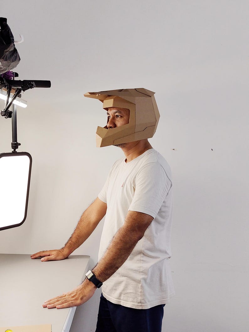 May include: A tan cardboard helmet with a geometric, angular design featuring a prominent visor and a wide face opening. The helmet is constructed from folded paperboard panels, showcasing a structured, faceted appearance that covers the head.