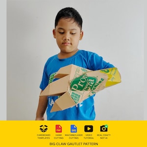 May include: A cardboard gauntlet featuring a large claw design, constructed from recycled packaging materials with visible green and yellow branding text. The structure includes pointed finger extensions and a hollow sleeve for the arm.