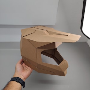 May include: A geometric helmet constructed from brown cardboard, featuring a prominent visor and a large open face area. The structure is composed of multiple angular panels joined together to form a full-coverage headpiece with a distinct motocross style.