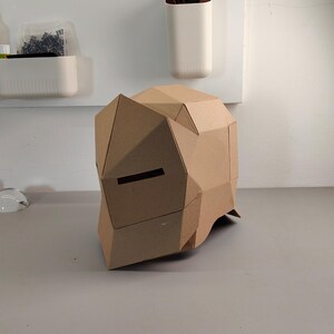 May include: A geometric cardboard helmet with a visor opening. The helmet is constructed from folded, brown cardboard pieces, creating a polygonal shape. The design suggests a DIY project or costume element.