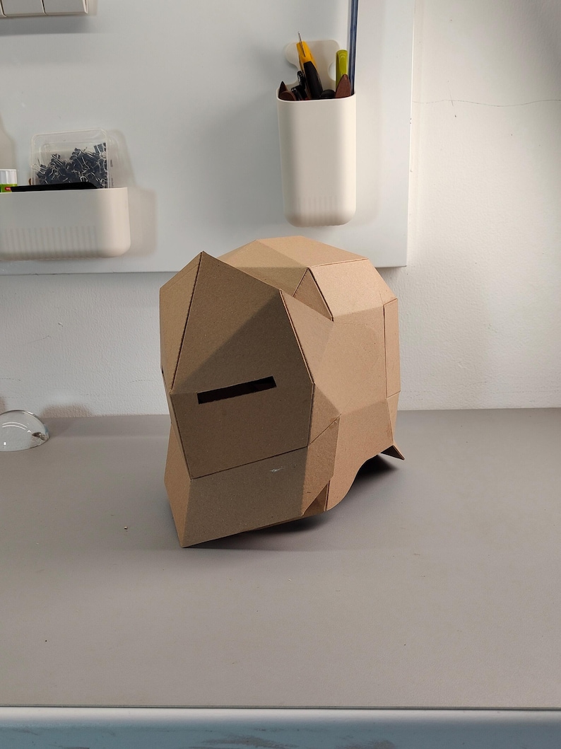 May include: A geometric cardboard helmet with a visor opening. The helmet is constructed from folded, brown cardboard pieces, creating a polygonal shape. The design suggests a DIY project or costume element.