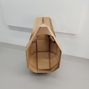 May include: A hollow, geometric mask constructed from tan corrugated cardboard. The structure features a series of interconnected, angular panels forming a faceted, helmet-like shape with a pointed top and an open front, showcasing internal seams.