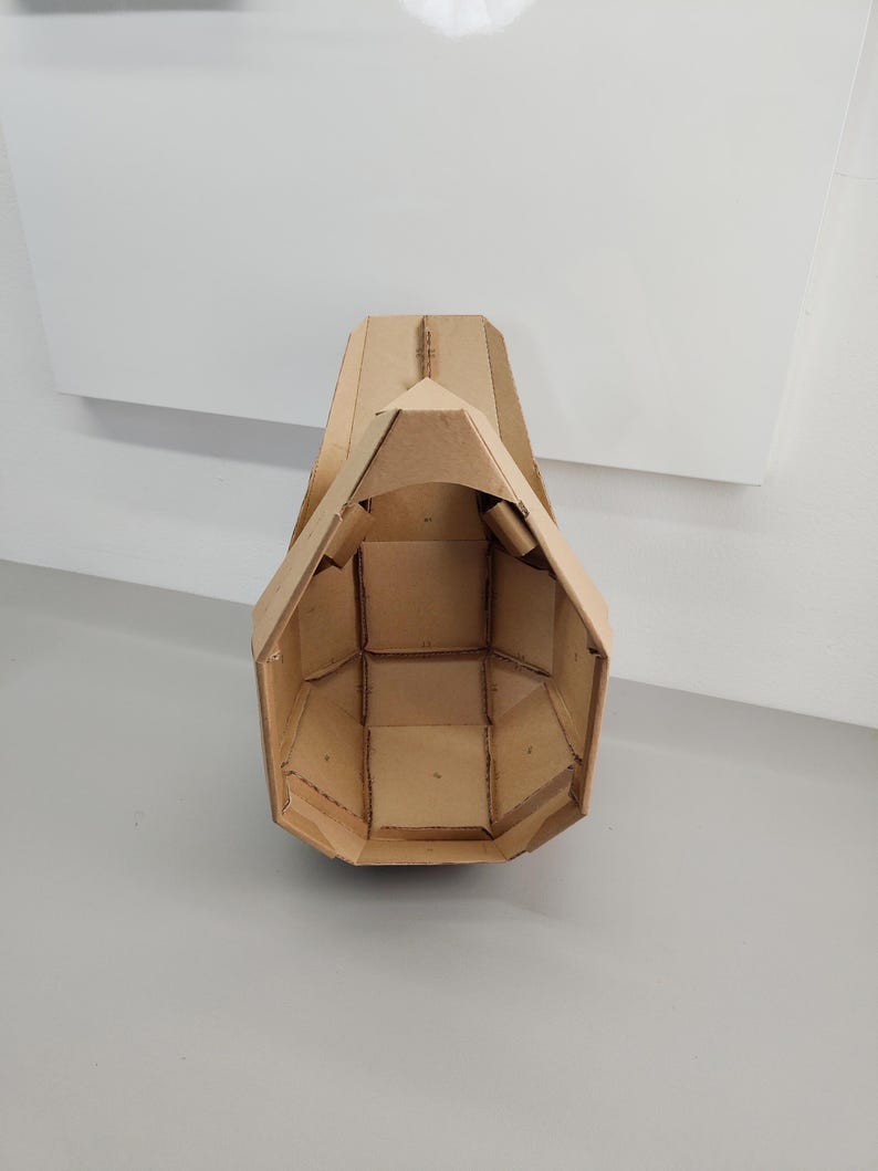 May include: A hollow, geometric mask constructed from tan corrugated cardboard. The structure features a series of interconnected, angular panels forming a faceted, helmet-like shape with a pointed top and an open front, showcasing internal seams.