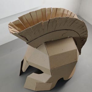 May include: A tan cardboard helmet featuring a prominent crest along the top. The helmet is constructed from multiple angular pieces of corrugated cardboard, forming a protective head covering with a wide opening for the face and eyes.