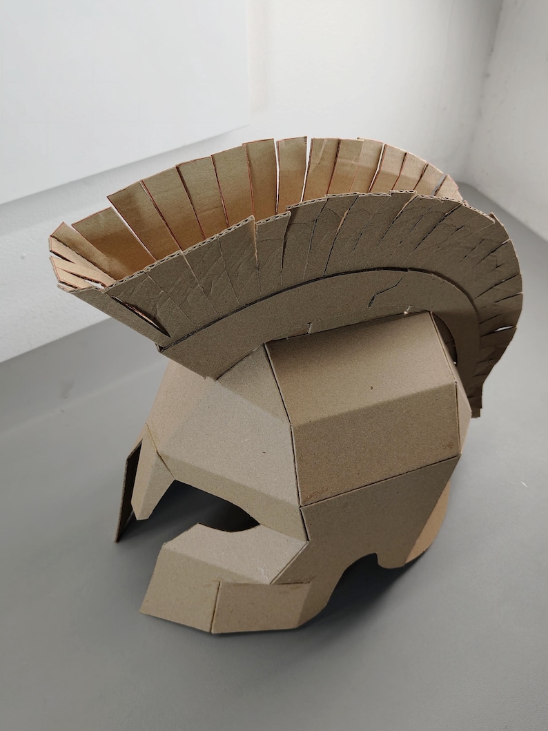 May include: A tan cardboard helmet featuring a prominent crest along the top. The helmet is constructed from multiple angular pieces of corrugated cardboard, forming a protective head covering with a wide opening for the face and eyes.