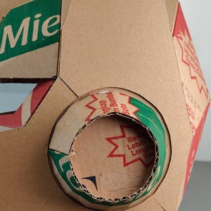 May include: A geometric structure constructed from brown cardboard packaging featuring printed text in green and red. The surface displays segments of product labels with the word Mie and various Indonesian phrases, including Bumbu Soto and Bawa Lebih Lengkap.