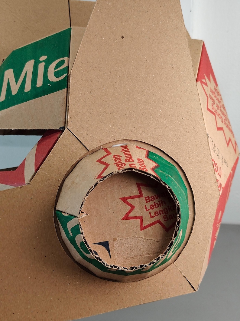 May include: A geometric structure constructed from brown cardboard packaging featuring printed text in green and red. The surface displays segments of product labels with the word Mie and various Indonesian phrases, including Bumbu Soto and Bawa Lebih Lengkap.