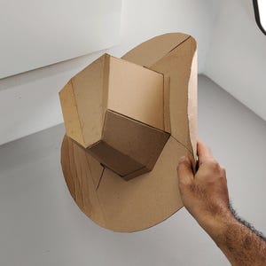 May include: A geometric cardboard sculpture with a layered design, featuring an angular central structure and a curved base. The sculpture is held by a hand, highlighting its form and construction. The cardboard is a natural brown colour.