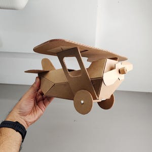 May include: A model aeroplane constructed from brown corrugated cardboard. The biplane features a top wing, a fuselage with angular sides, a front propeller, and two circular wheels attached to the sides. The structure is held together with visible joints.