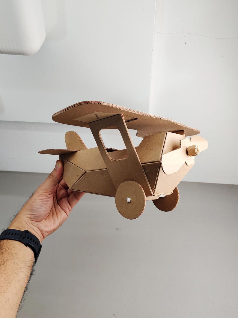 May include: A model aeroplane constructed from brown corrugated cardboard. The biplane features a top wing, a fuselage with angular sides, a front propeller, and two circular wheels attached to the sides. The structure is held together with visible joints.