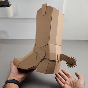 Puede incluir: Una escultura tridimensional de bota vaquera construida con cart&oacute;n de color marr&oacute;n claro. La bota presenta una ca&ntilde;a alta, una punta afilada y una espuela decorativa unida al tal&oacute;n con una rodaja en forma de estrella, todo hecho de cart&oacute;n plegado.