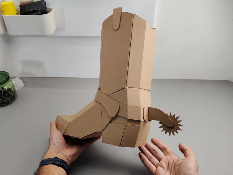 Puede incluir: Una escultura tridimensional de bota vaquera construida con cart&oacute;n de color marr&oacute;n claro. La bota presenta una ca&ntilde;a alta, una punta afilada y una espuela decorativa unida al tal&oacute;n con una rodaja en forma de estrella, todo hecho de cart&oacute;n plegado.