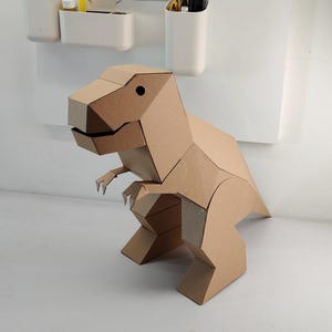 May include: A geometric dinosaur figure constructed from light brown cardboard. The sculpture features a blocky, angular design with a large head, small arms, and a thick tail, showcasing a clean, folded paper aesthetic with a simple black dot eye.